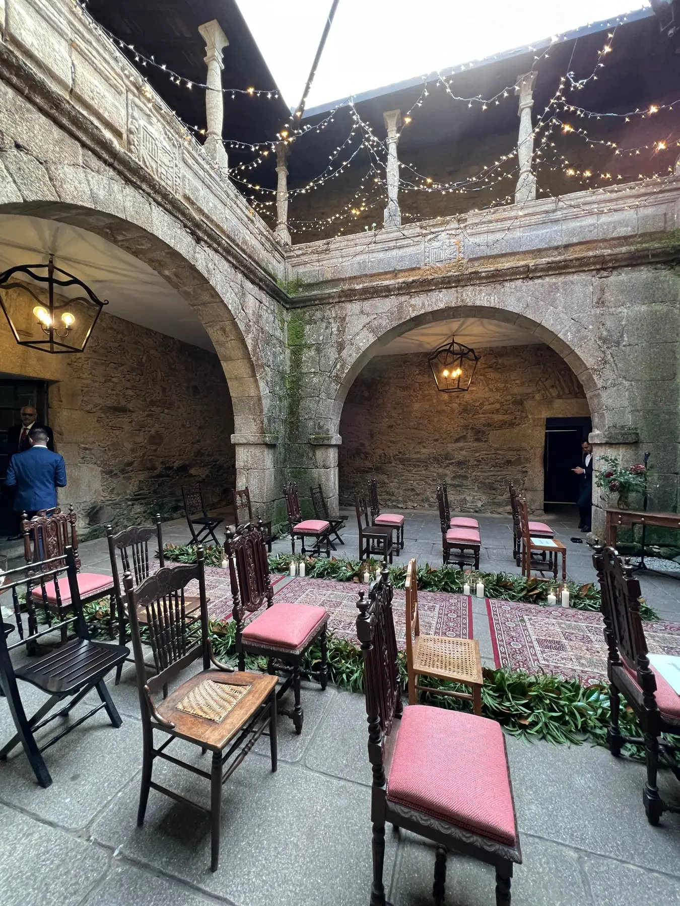 Sillas de madera con cojines rosas y alfombras en el claustro, bajo arcos de piedra y guirnaldas de luces, montadas para una ceremonia