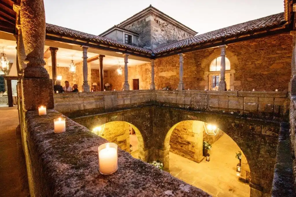 El claustro con velas sobre el pretil, columnas de piedra, arcos iluminados y una sala con invitados al fondo
