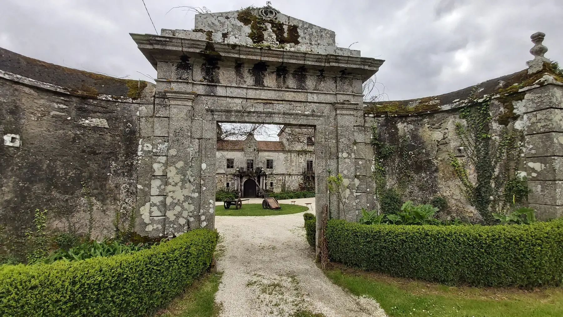 El portalón de piedra con hiedra, un camino de grava y setos de boj, y el pazo al fondo