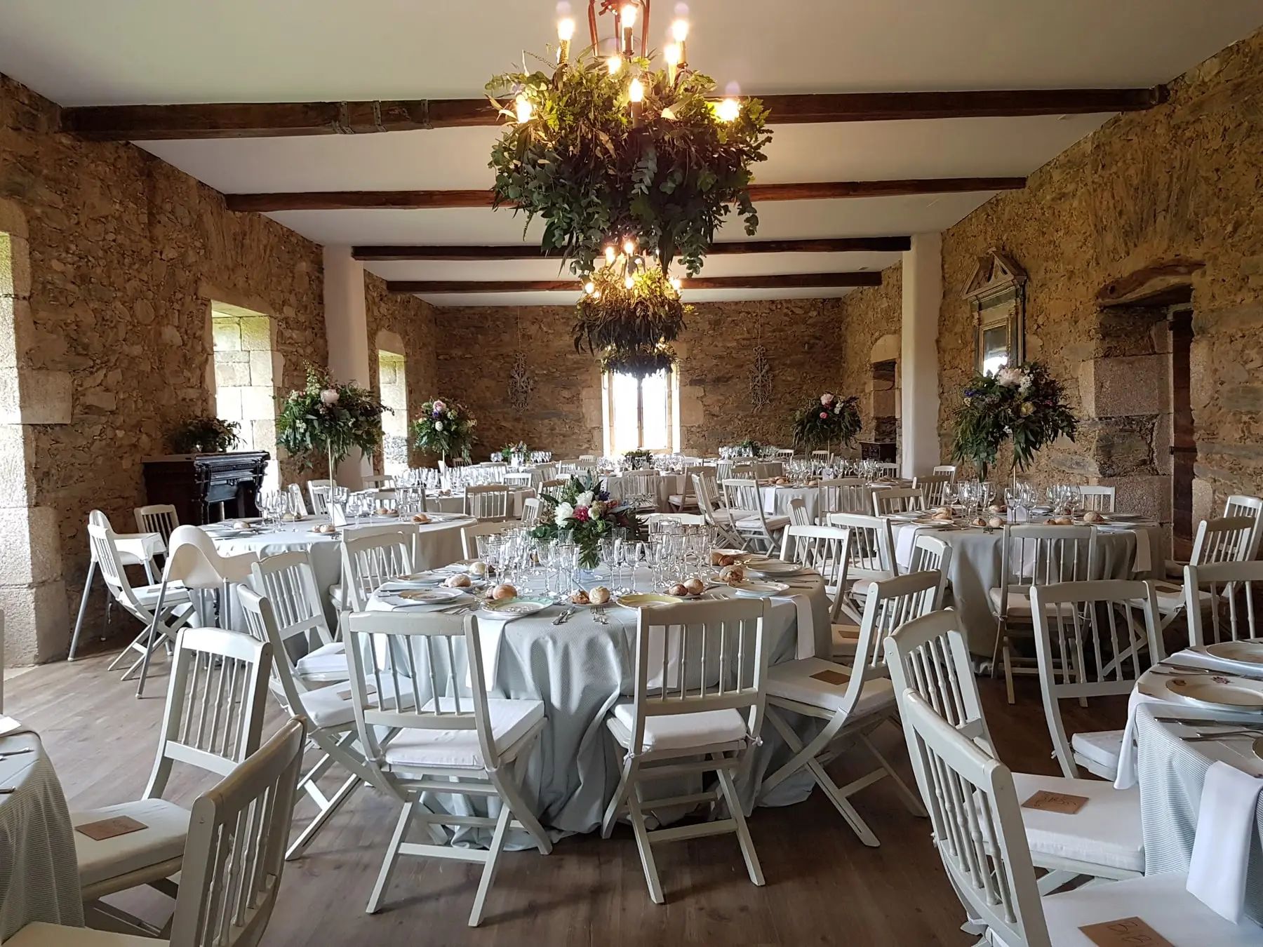 El salón principal montado para un banquete: mesas redondas, sillas blancas, vigas de madera y una gran lámpara de flores