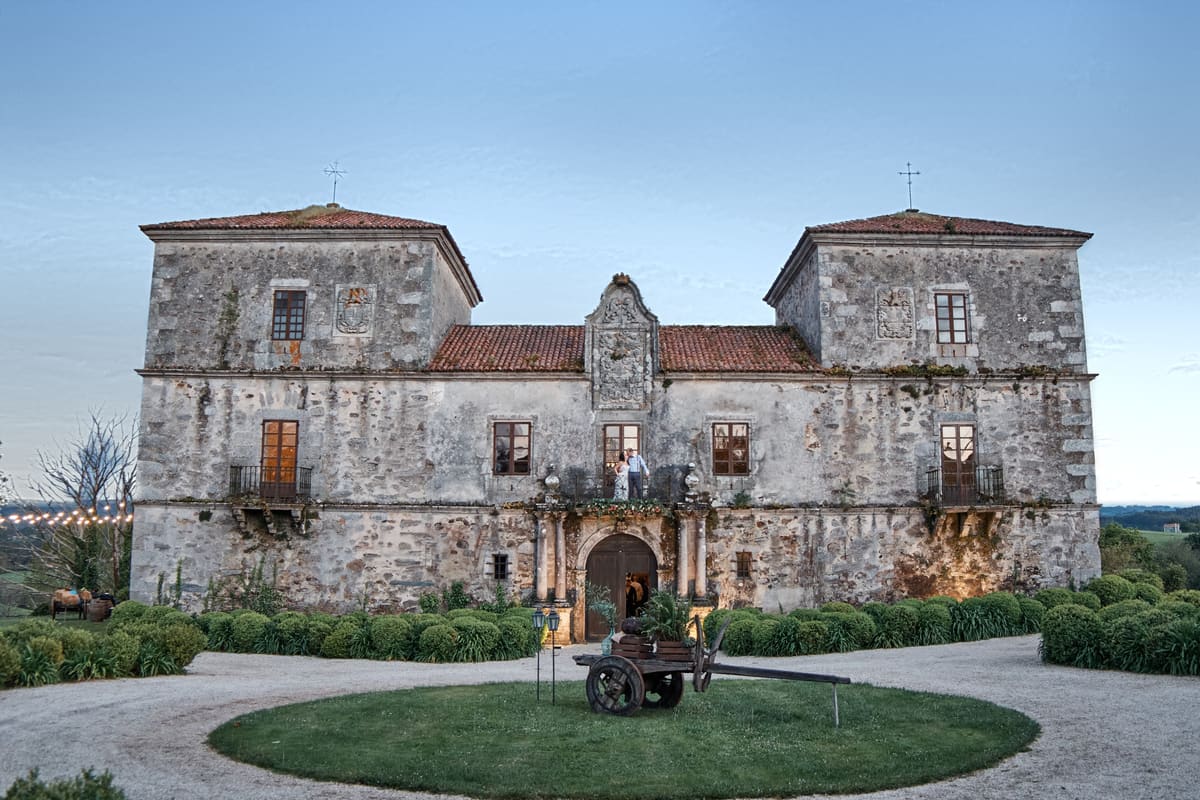 El Pazo de San Tirso al caer la tarde: la fachada de piedra con dos torres, el escudo sobre el balcón central, setos de boj y una carreta sobre el césped